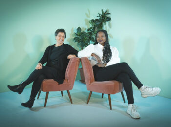Das Foto zeigt Monika Heinold und Aminata Touré. Beide sitzen entspannt lächelnd nebeneinander auf zwei Sesseln. Im Hintergrund ist eine grün angestrahlte Wand zu sehen.