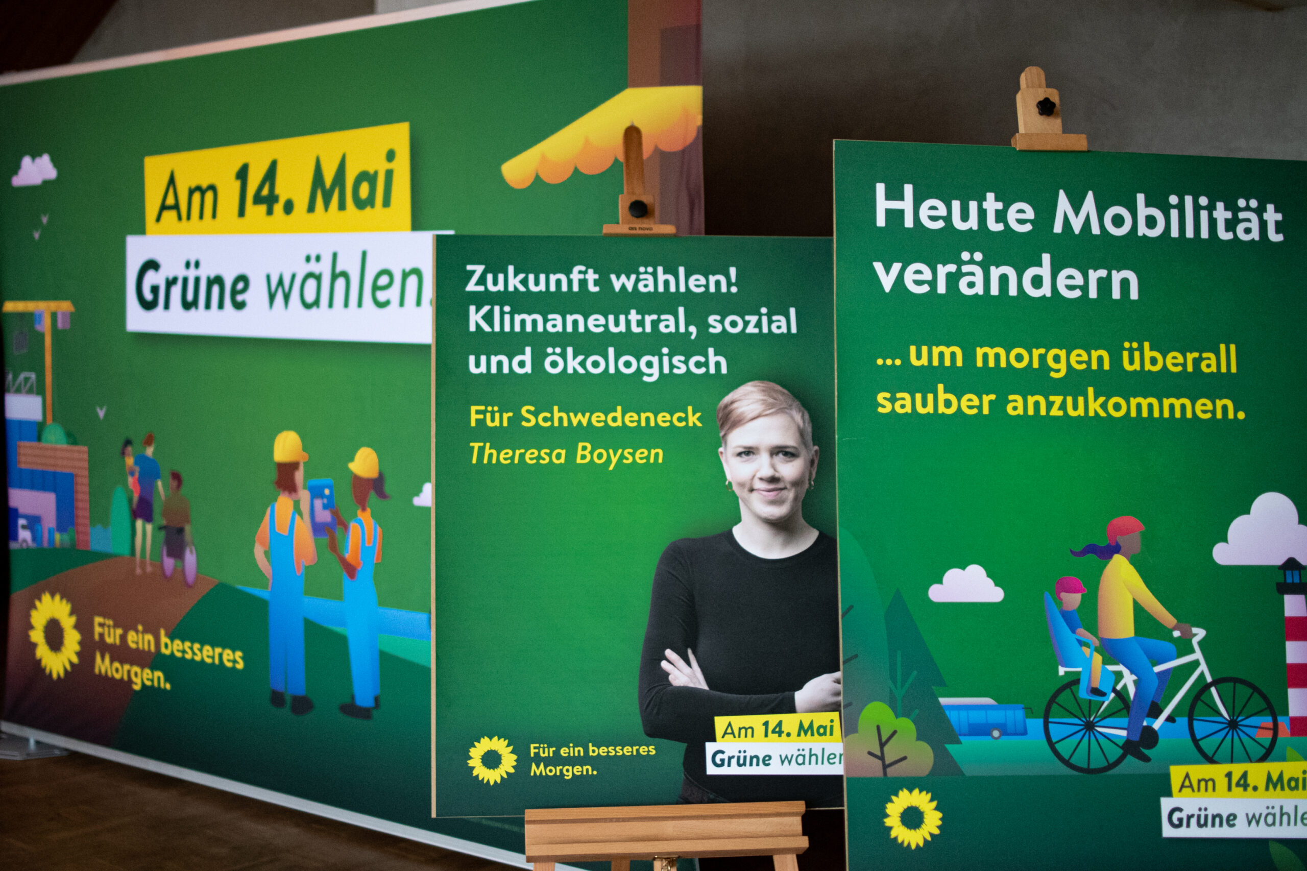 Ein Grünes Plakat zum Thema Mobilität und ein Kandidatinnenplakat stehen vor dem Großflächenmotiv mit dem Titel "Am 14. Mai Grüne wählen!"